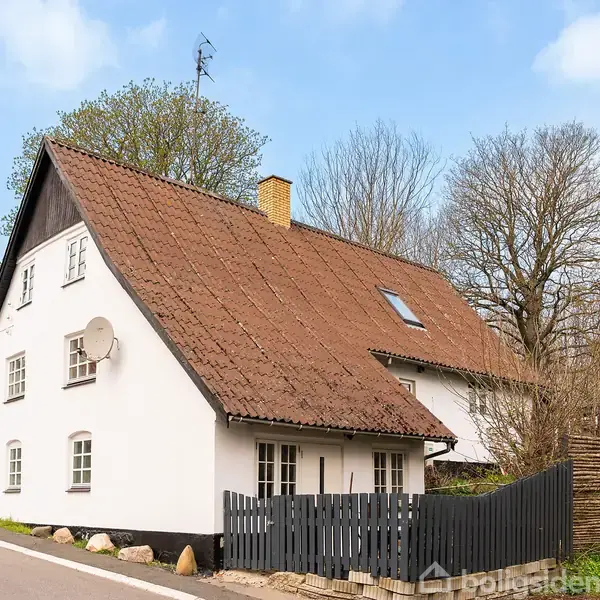 Hvidt hus med rødt tegltag har parabolantenne på siden. Står ved landevej, omgivet af træer og hegn. Den blå himmel ses i baggrunden.