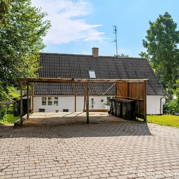 Carport står tom på en brostensbelagt indkørsel ved siden af et hvidt hus med mørkt sadeltag. Grønt buskads og træer omkranser området.