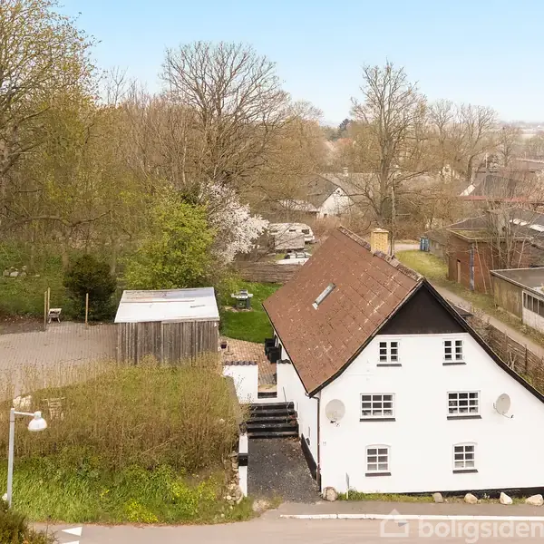 Et hvidt, to-etagers hus med brun tag ligger i et grønt områdemed træer omkring. Der er en parkeringsplads ved siden af huset. Baggrunden viser flere bygninger og åbne marker.
