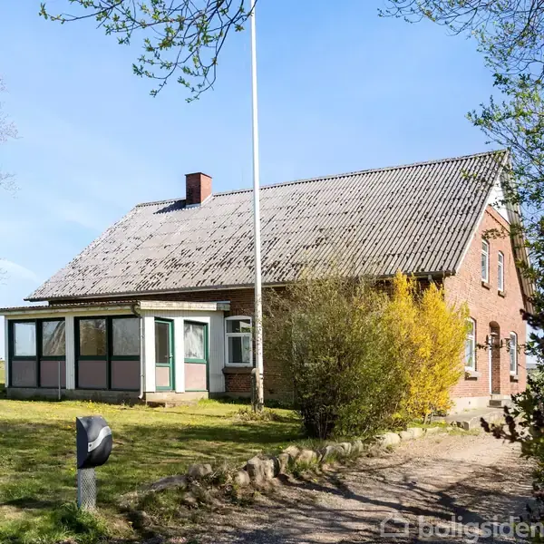 Et rødt murstenshus står stille på en grusvej. Det har et skråtag med nogle mospletter. Omgivet af grønne omgivelser og træer under en klar, blå himmel.