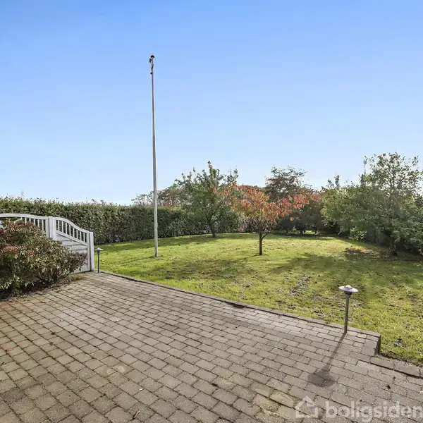 Fliseterrasse fører ud til en græsplæne med enkelte træer og en flagstang. Omkring plænen er der høje grønne hække under en klar blå himmel.