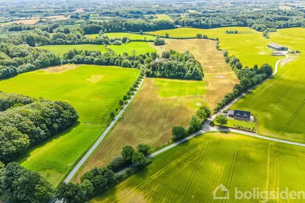 Landejendom omgivet af grønne marker og skove. Ejendommen omfatter en stor gul mark med omliggende veje og en enkelt bygning i højre forgrund. Landskabet er frodigt og landskabeligt.