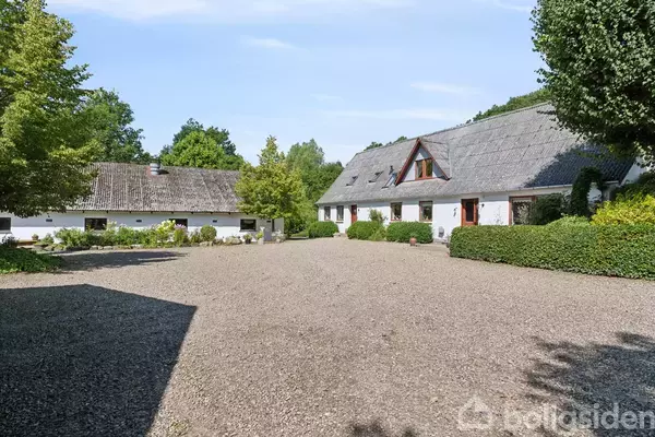To hvidkalkede bygninger med stråtag står i en solrig gårdsplads omkranset af grønne buske og træer, under en klar blå himmel.