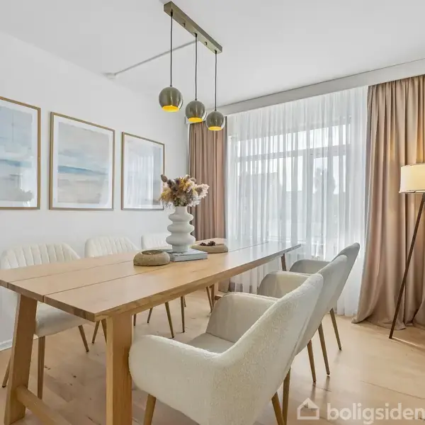 Et spisebord med seks beige stole står i et lyst rum. Over bordet hænger tre lamper. Rummet har lyse gardiner, tre billeder på væggen, og en gulvlampe i hjørnet.