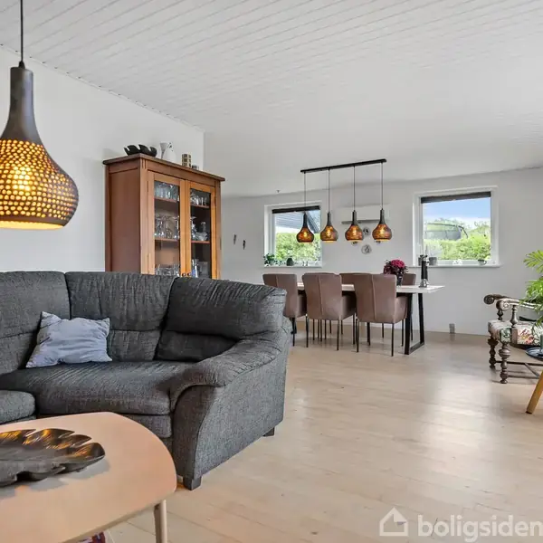 Grå sofa står i en lys stue med trægulv, hvor en sort lampeskærm hænger over sofabordet. Bagved ses spisebord og stole ved vinduer. Plantestander i hjørnet.