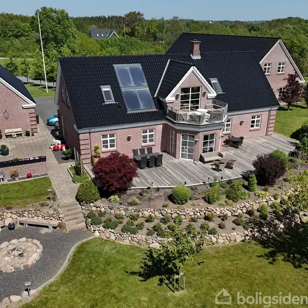 Stort murstenshus med solpaneler på taget, omkranset af en velplejet have med stensætninger og græsplæner. Ved siden af ses en mindre bygning, placeret i et grønt, landligt område.