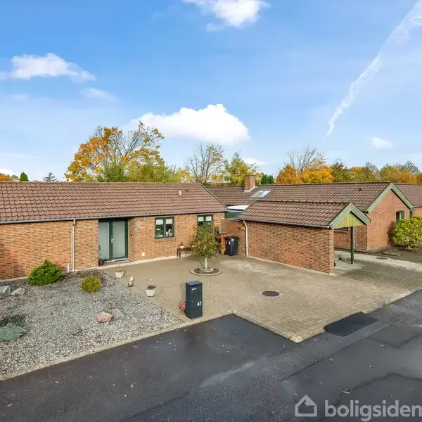 Rækkehuse med røde mursten og rødt tag ligger langs en stille vej. Nogle huse har små forhave med grus og buske. Bagved ses træer med efterårsfarver under en blå himmel.