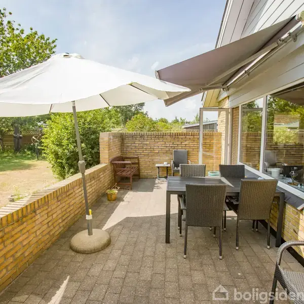 En terrasse med et bord og seks stole står under en stor parasol. Terrasseområdet er omgivet af en lav mur, med en have i baggrunden og en markise over vinduerne.