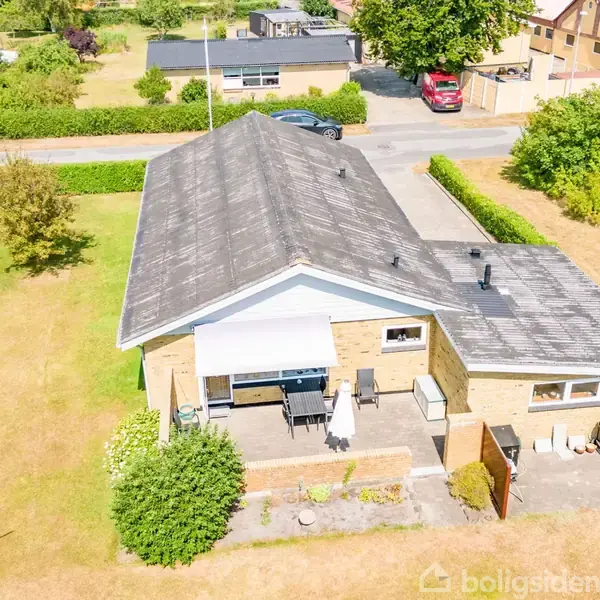 Hus set fra oven, omgivet af en stor have med fliser og græsplæne. Terrasse med havemøbler og parasol. Beliggende i en rolig villavej med flere huse i baggrunden.