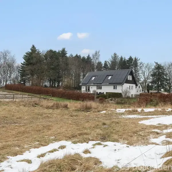 Hvidt hus med sort tag står stille i et åbent landskab, omgivet af træer og marker med spredt sne.
