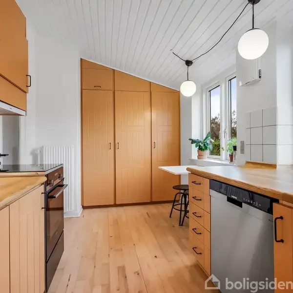Køkken med træskabe står stille i et lyst rum. Bordpladerne er også af træ. Rummet har hvidt loft og vægge, med store vinduer, der giver naturligt lys.