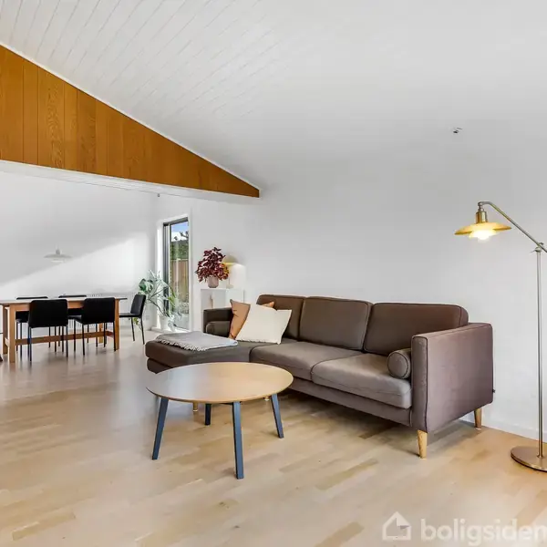 En brun sofa står i en lys stue under et skråt loft. Et rundt sofabord står foran sofaen. Til venstre ses et spisebord med stole ved en hvid væg.