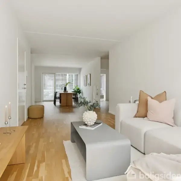 Hvid sofa med puder står i en stue med lyst trægulv. Et gråt sofabord foran sofaen. En plakat til venstre viser abstrakt kunst. I baggrunden ses et bord nær vinduet.
