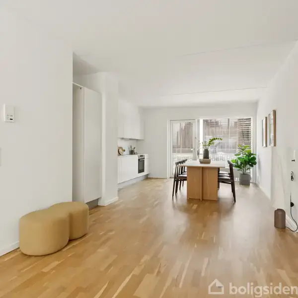 Et lyst, moderne køkken-alrum med et spisebord og stole i midten, omgivet af lyse trægulve. Store vinduer i baggrunden slipper naturligt lys ind.
