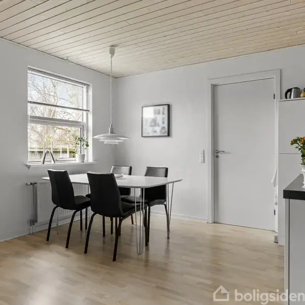 Et spisebord med fire sorte stole står i et lyst køkken-alrum med træloft og stort vindue. En hvid lampe hænger over bordet, og en plante står på en skænk.