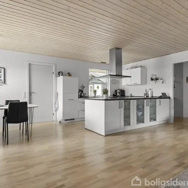 Køkken med hvidt og sort møblement, komfur og emhætte; i baggrunden ses et spisebord med stole. Lys trægulv og loft. Lysindfald gennem vinduer, enkel indretning.