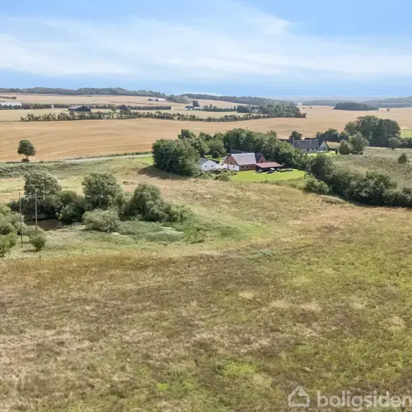 En gård ligger omgivet af marker og træer på en stor eng. I baggrunden ses bølgende landskaber og flere skove.