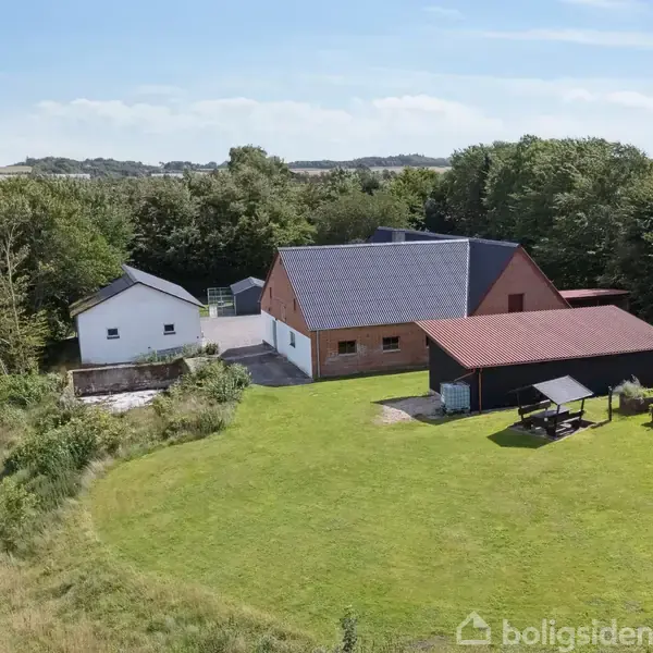Hus med rødt tag ligger i en grøn have med havemøbler nær et skovklædt område. En mindre, hvid bygning står til venstre, omgivet af marker og træer under en blå himmel.