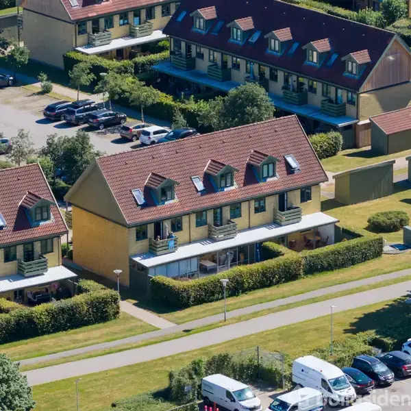 Rækkehusene med røde tage står i en række. Der er parkerede biler foran dem, omgivet af grønne områder og træer i en bebygget forstad.