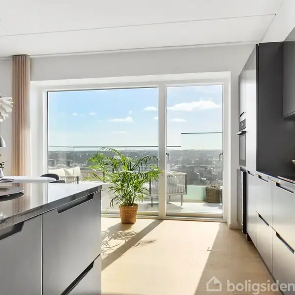 Køkken med moderne grå skabe beliggende langs væggene. Et stort vindue åbner udsigten til en balkon med en plante, mens sollys fylder rummet.