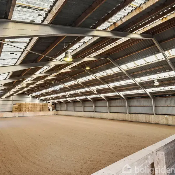 En stor ridehal med sandbund, oplyst af loftslamper. Omgivet af metalvægge og tagvinduer, som skaber en lys atmosfære.