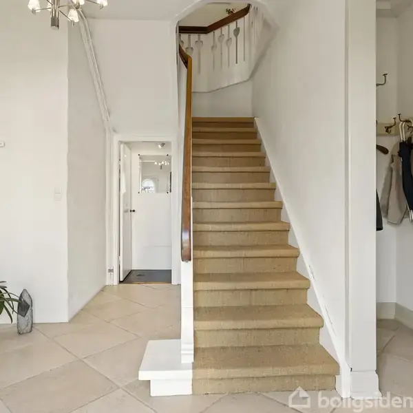 En trætrappe fører opad i en lys entré med lysekroner og planter. Jakker hænger på knager ved siden af trappen.