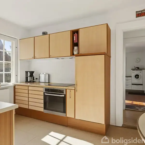 Køkken indeholder lyse træskabe og ovn, sollys strømmer ind gennem store vinduer. I baggrunden ses en vaskemaskine. På væggen hænger skiltet: “What part of ‘Thou shall not!’ didn’t you understand?”