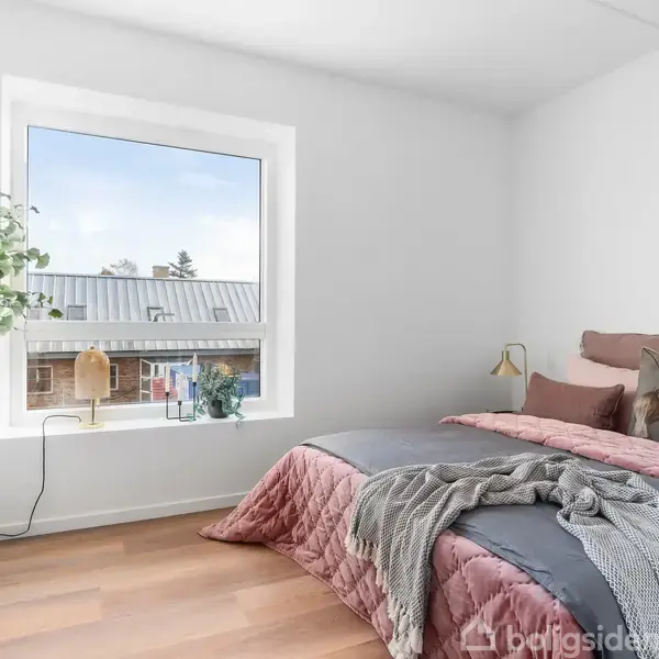 Et soveværelse med en redt seng med puder og sengetæppe, lille vindue med udsigt til bygninger, samt planter og lamper i lyse og moderne omgivelser.