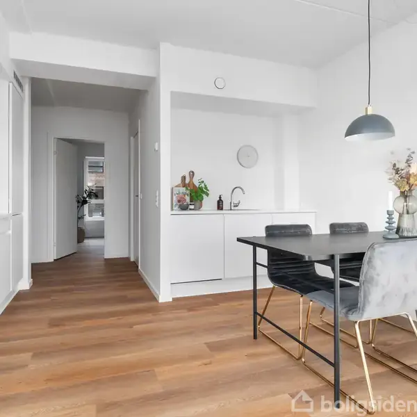 Et moderne køkken-alrum med træparketgulv; et sort spisebord med fire grå stole, en loftslampe over bordet, og en vase med blomster. Køkkenet har hvide skabe og integreret ovn.