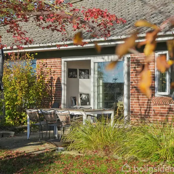 Rødt murstenshus med udendørs terrasse, hvor havebord og stole står opstillet. Omgivet af efterårsløv og grøn beplantning i en solrig have.
