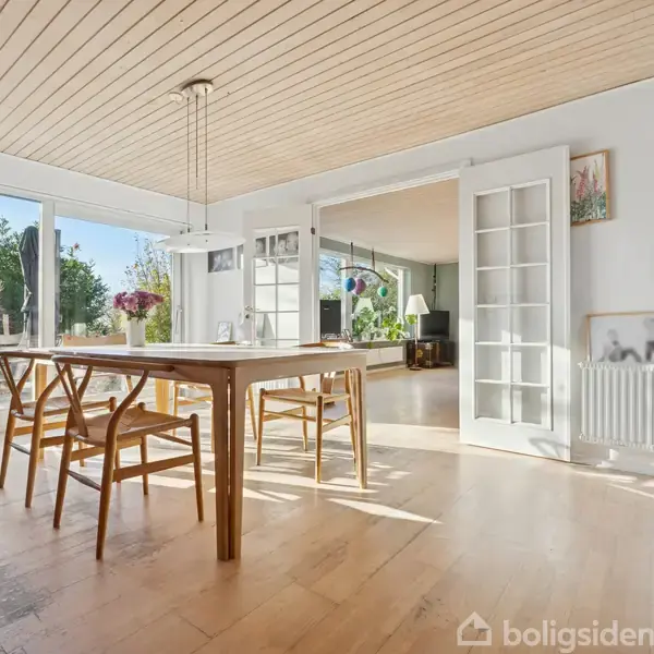 Spisebord står i et lyst rum med træloft, omgivet af fire stole. Vinduer og glasdøre til højre giver udsigt til terrasse og have. Vægdekorationer ses til højre.