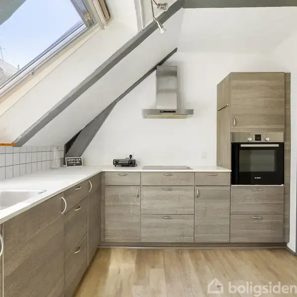 Køkken, indrettet med moderne træskabe og indbyggede apparater, under skrå loftsvinduer, der lader lys strømme ind. Lyse trægulve og hvid flisebagvæg skaber et lyst rum.