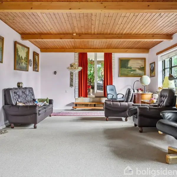 Stue med sorte lænestole og sofaer, træloft og tæppebelagt gulv. Væggene har malerier, og der er store vinduer med røde gardiner i baggrunden.