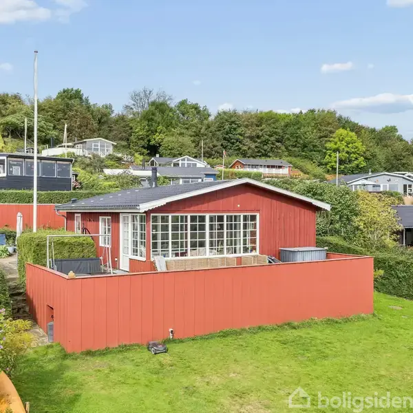 Rødt træhus omgivet af grønne hække og græsplæne, står på en solrig dag med et bagtæppe af flere huse og træer på en bakketop.