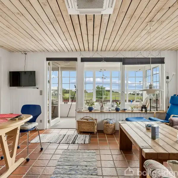 Stue med træloft og terracotta-flisegulv. Sofa og sofabord i højre side, spisebord med stole til venstre. Store vinduer viser grønt landskab udenfor. Lys og indbydende atmosfære.