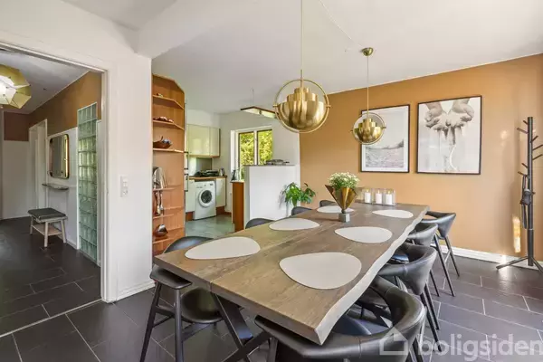 Et spisebord med sorte stole er dækket med lyse dækkeservietter. To gyldne lamper hænger over bordet. Rummet har orangerøde vægge og kunstbilleder. Køkken ses i baggrunden.