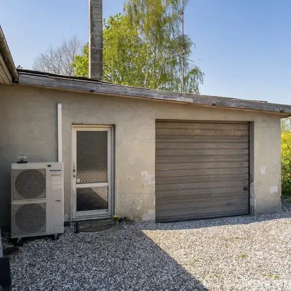 Bygning med gråt facade monteret med en varmepumpe ved siden af en glasdør. En garageport til højre. Omgivelserne består af grus og grønne buske under en klar blå himmel.