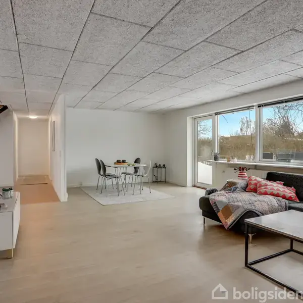 En lys stue med en sofa og sofabord, vender mod store vinduer. Et spisebord med stole står i baggrunden nær en hvid væg.