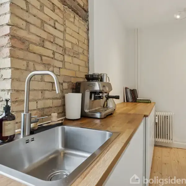 Vask med vandhane i køkken med træbordplade. Kaffe- og håndaftappemaskiner står ved siden af. Muren er delvist blottet. Grønne planter ses i vindueskarmen. Væggene er hvide.