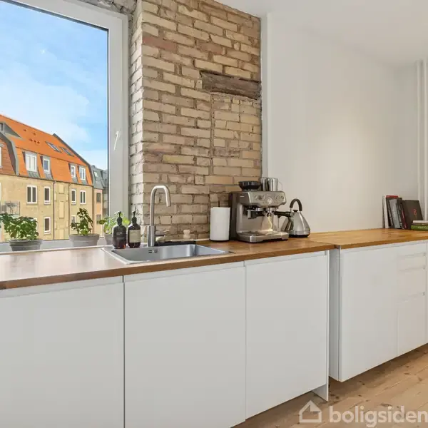 Køkkenbord af træ med hvidt skab oppe imod en murstensvæg; en kaffemaskine står på bordet. Through vinduet ses ældre bygninger med røde tage og blå himmel udenfor.