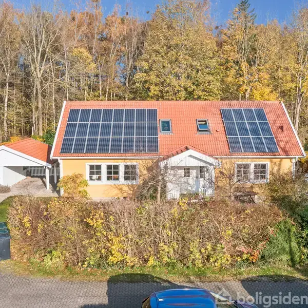 Et gult hus med rødt tegltag og solpaneler står i en skovklædt område. En bil holder i indkørslen ved siden af en garage.