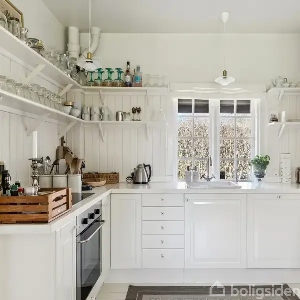 Et hvidt køkken med åbne hylder fyldt med glas og flasker. En kogeplade og ovn er indbygget i kabinettet. Ved vinduet står en elkedel og planter.