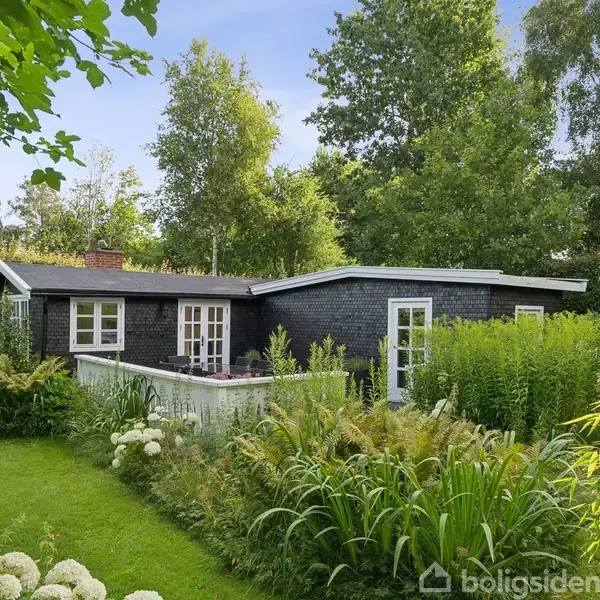 Sort sommerhus med hvidrammede vinduer står stille omgivet af frodig have med grøn plæne, buske og træer under en blå himmel.