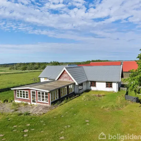 En landlig ejendom med en hovedbygning og tilbygning, omkranset af græsarealer. En klar blå himmel og et stort træ til højre danner en fredelig atmosfære.