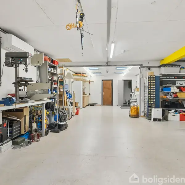Et værksted er organiseret med værktøjer hængende på væggen til venstre og en række hylder fyldt med udstyr til højre. Gulvet er rent og ryddeligt.
