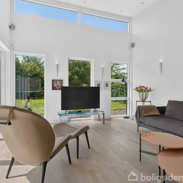 Et moderne stue med en sofa og lænestole, hvor en fladskærms-TV står på et lavt bord. Store vinduer giver udsigt til en grøn have, dekoreret med kunstværker og blomster.