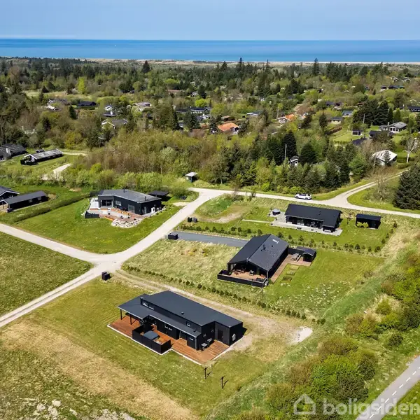 Moderne enfamiliehuse ligger langs grusveje i et grønt villakvarter tæt på skov og kyst. Havet ses i baggrunden, mens spredte træer og plæner præger landskabet.