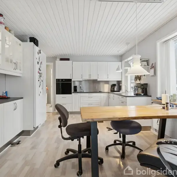 Et køkken med hvide skabe og træbord, to sorte kontorstole ved bordet, moderne apparater og store vinduer, der giver udsigt til sneklædt have.