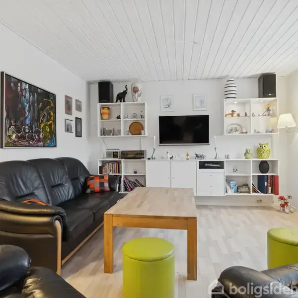 Sorte lædersofaer omkring et træbord, stående på et lyst trægulv. Fjernsynet er monteret på en hvid reol med dekorationer. Vindue til højre bringer lys ind.