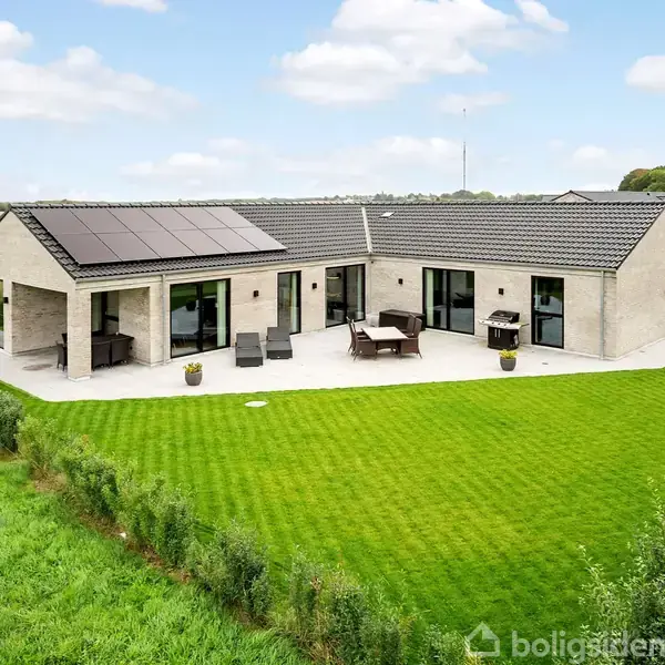 Et moderne hus med solpaneler på taget står stille på en stor, velplejet græsplæne. Omkring huset er der buske, og det ligger i et åbent, landligt område.